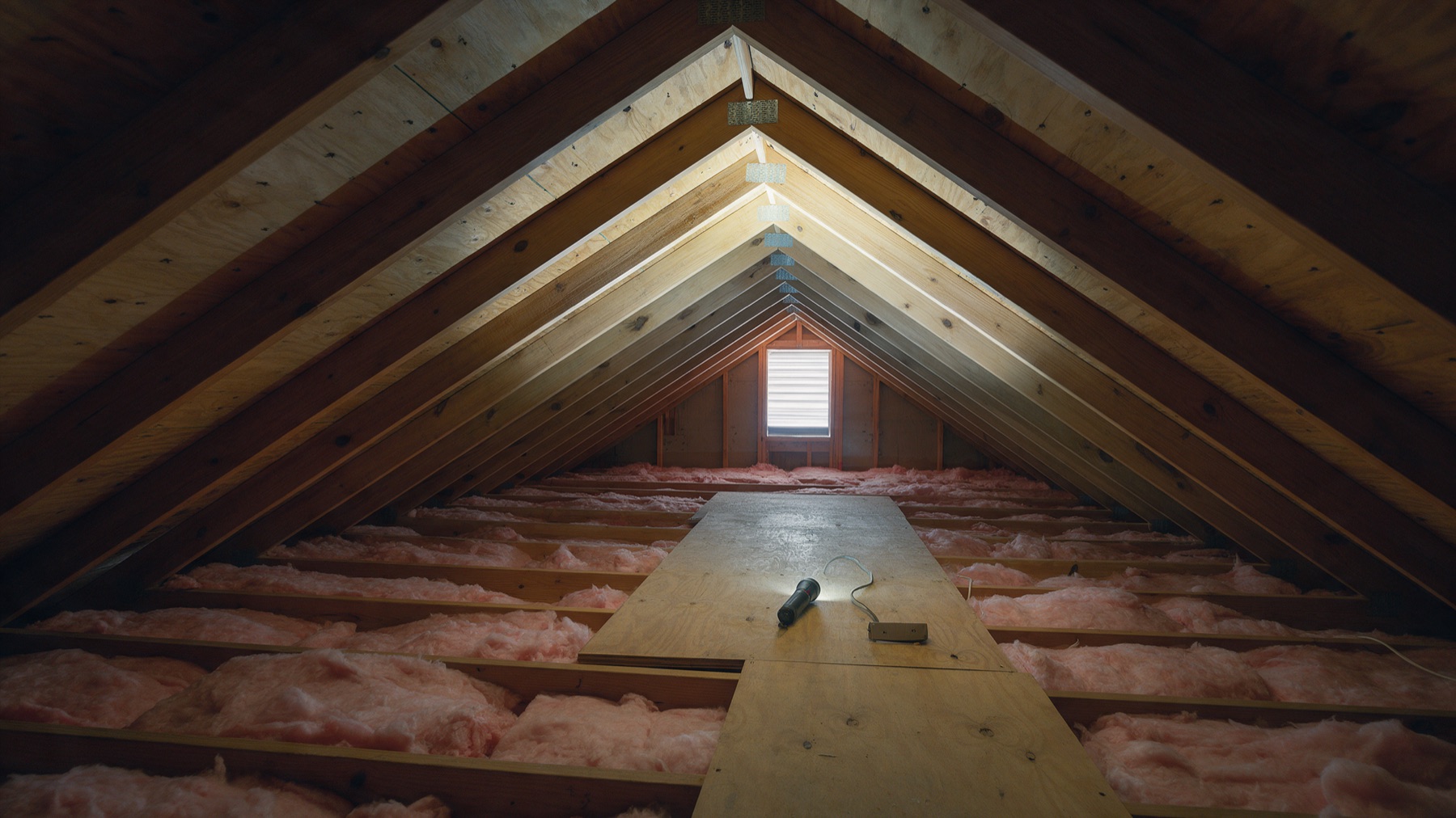 The inside of a tidy residential attic looking toward a gable vent, with exposed wood rafters, pink fiberglass insulation between the joists, and a flashlight resting on a plywood walkway board.