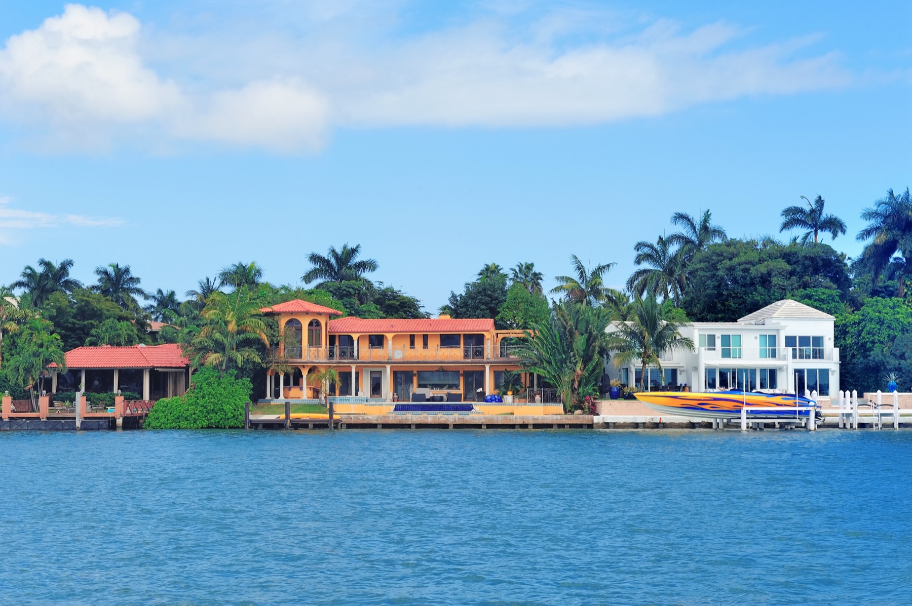 Waterfront homes in Miami seen from across the water, a terracotta-roofed Mediterranean-style house and a red-tiled pavilion among tall palms beside a white modern house, under a blue sky with clouds.