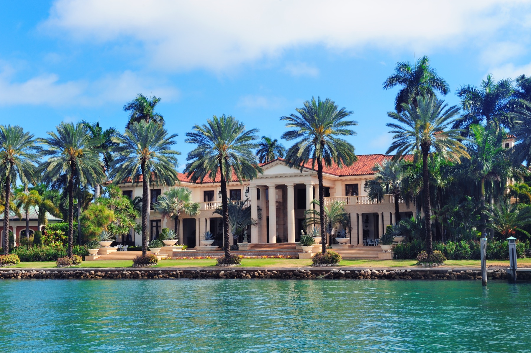 A large cream-colored house with white columns and a terracotta tile roof among tall palm trees, seen across calm turquoise water on a sunny day in Miami.
