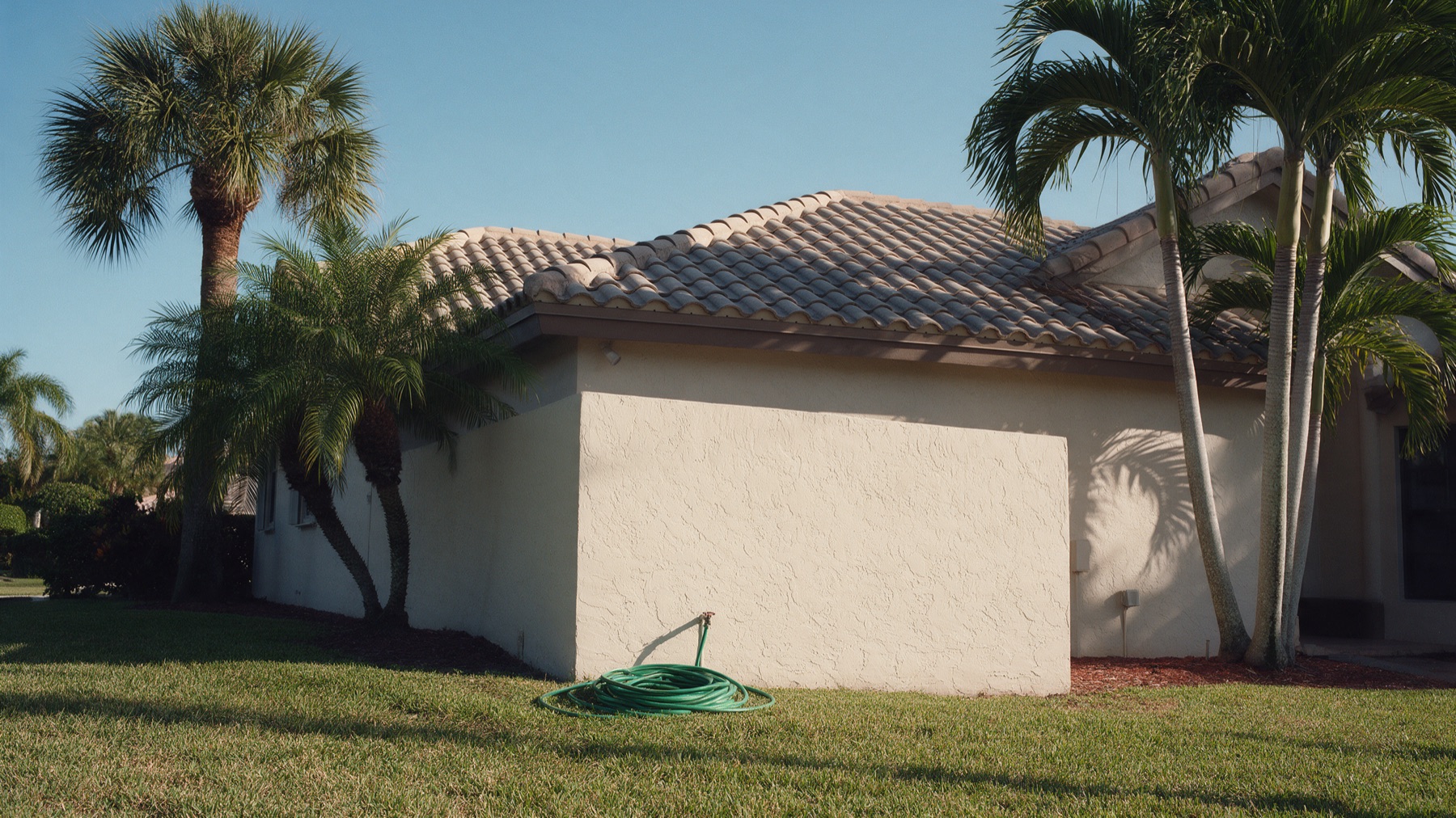 A green garden hose coiled on a lawn beside a white stucco house with a tile roof and palm trees under a clear blue South Florida sky.