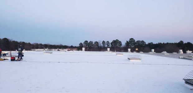 Snow across a flat commercial roof, bare tree line on the horizon.