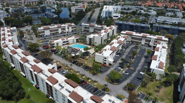 Aerial over a garden-apartment community — pool deck, parking and completed roofs in one frame.